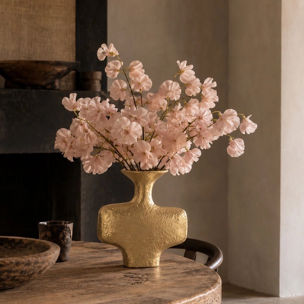 Everley Metal Vase in textured gold finish filled with soft pink flowers, displayed on a rustic wooden dining table in a warm neutral interior.