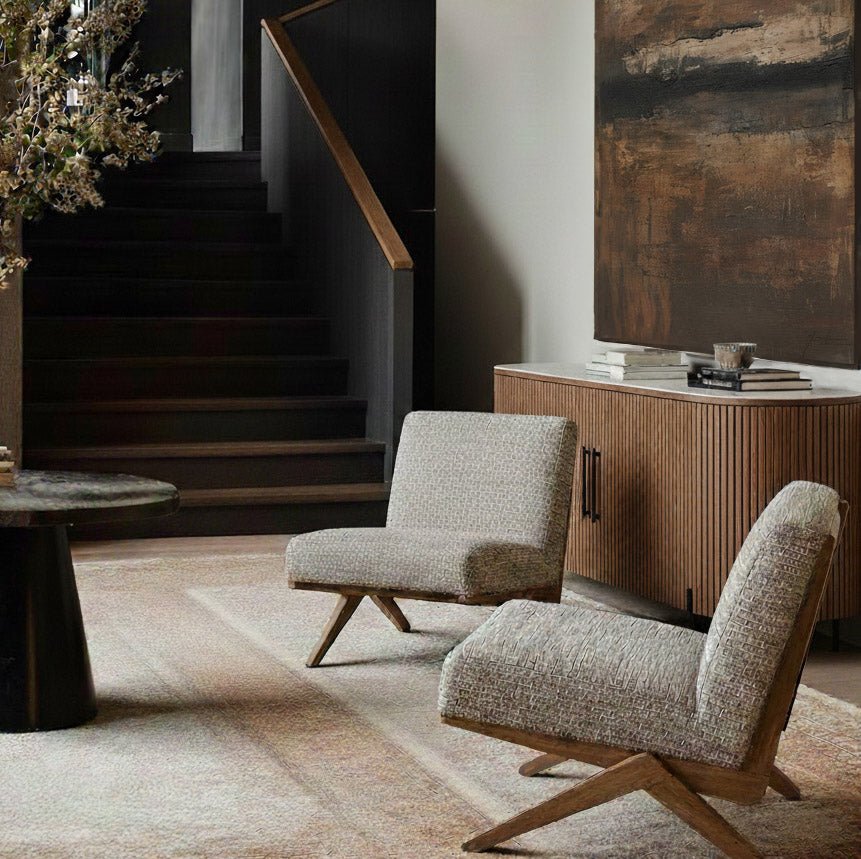 Abigail Ahern Exbury Modern Armchairs in soft grey patterned upholstery, featuring low angled silhouettes, wide cushioned seats and wooden legs, styled in a warm contemporary interior with a fluted wooden sideboard and dark staircase.