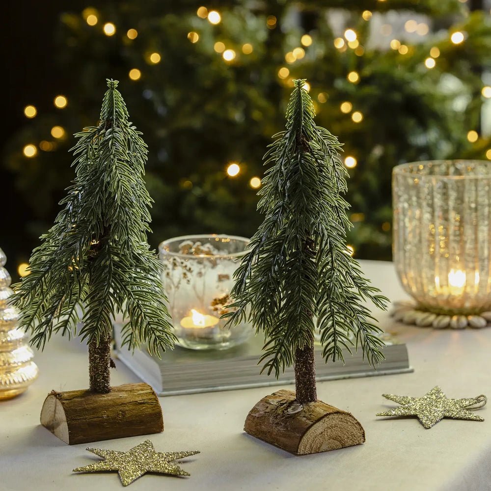Festive tree decorations with green branchlike foliage and dark trunks stand on curved wooden bases, arranged beside a patterned candleholder on a festive table.