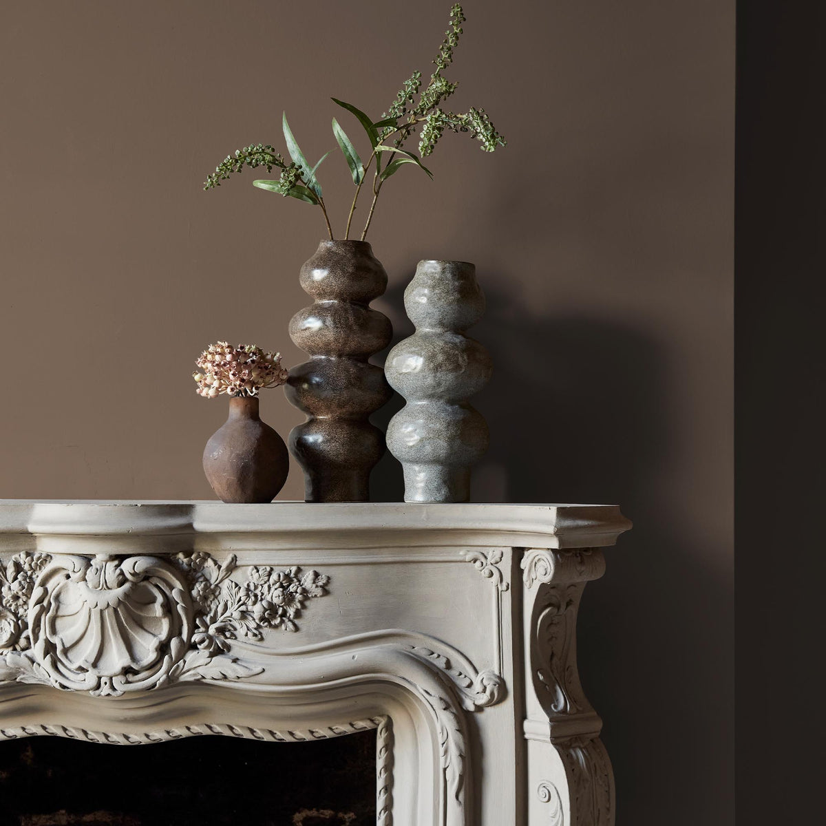 Three ceramic vases sitting on an intricate fireplace mantle in front of a wall in a soft brown paint colour.