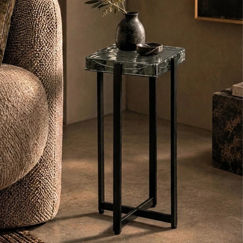 Fordham Glass Side Table features a square textured glass top and slim dark frame, styled beside a curved armchair with a vase and branches.