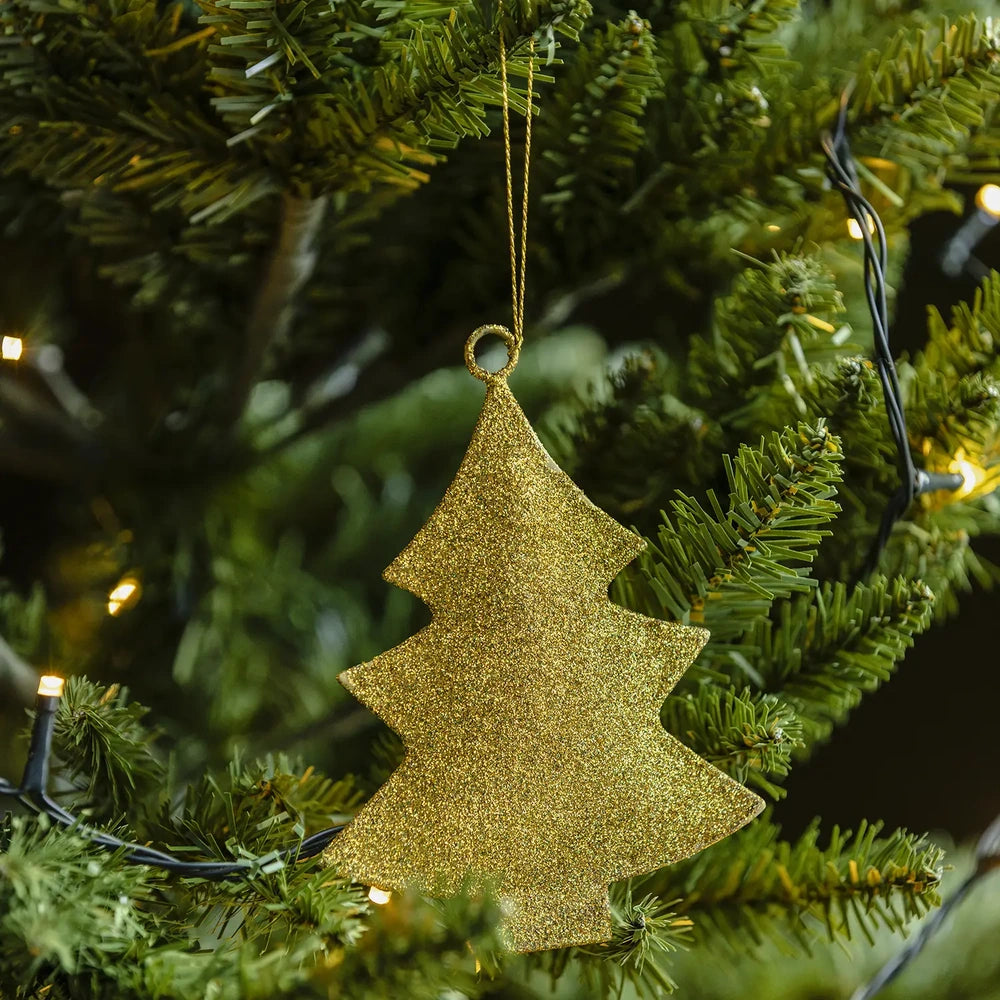 Glittering tree bauble shows a flat gold tree silhouette and densely sparkling surface in close view, suspended among green branches with warm lights behind.
