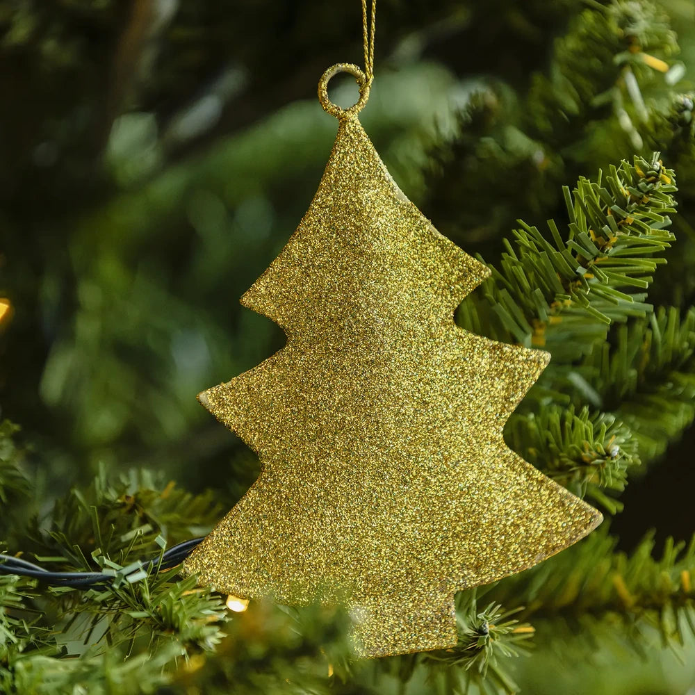 Glittering tree bauble hangs from a gold loop among green Christmas tree branches, showing its pointed outline and fine textured surface beside warm lights.
