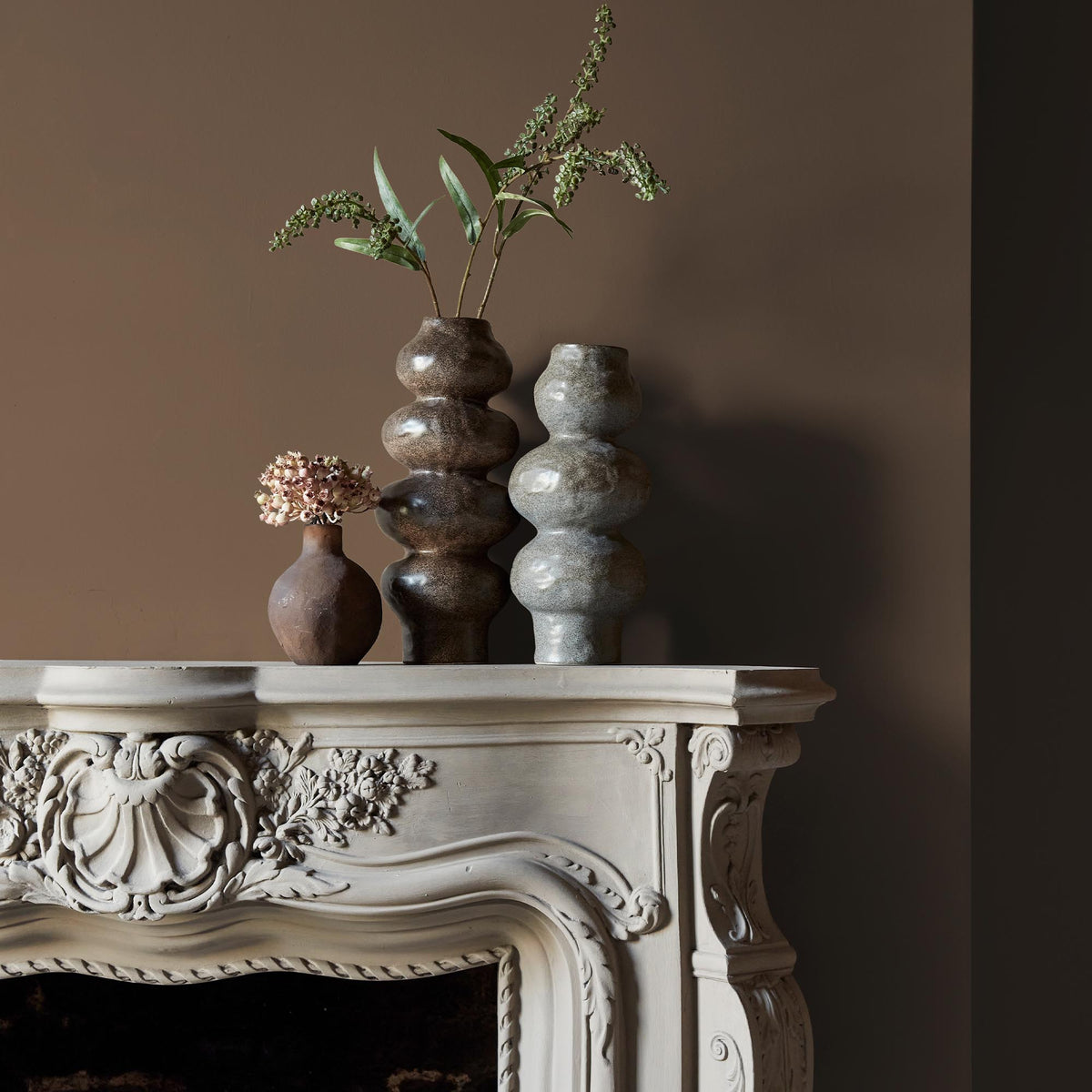 Three ceramic vases sitting on an intricate fireplace mantle in front of a wall in a warm brown paint colour with golden undertones.