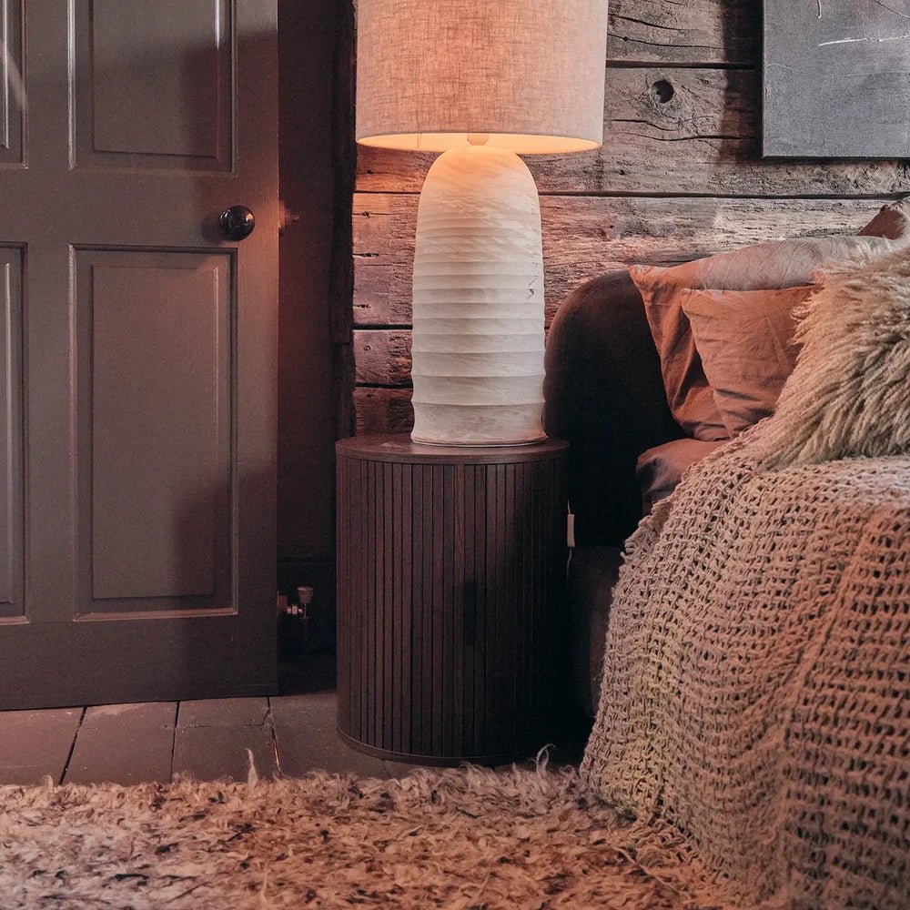 Halmore Wooden Bedside Table features dark vertical ribbing and a rounded form, positioned beneath a pale lamp beside a bed against rustic wooden panelling.