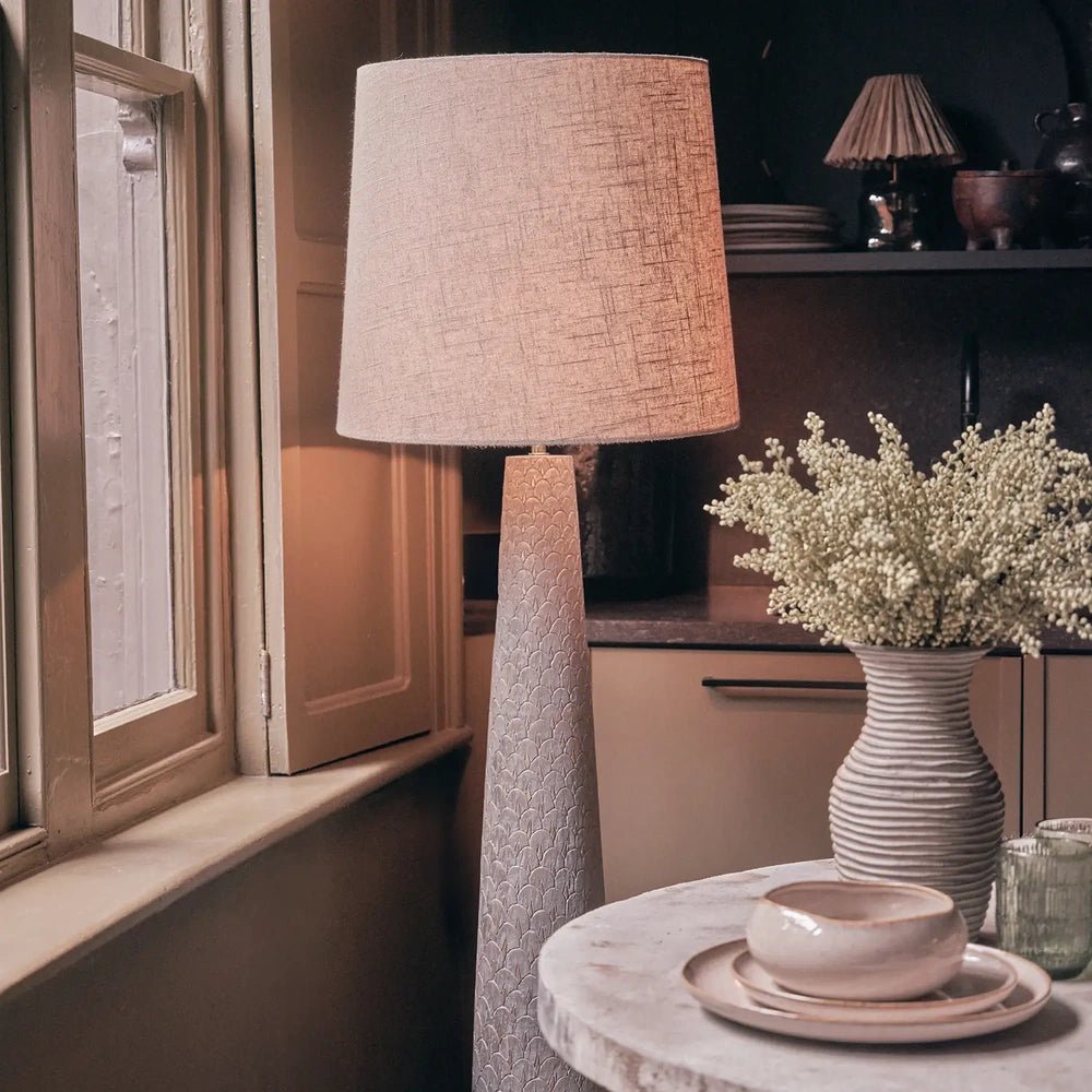 Haverford Polyresin Floor Lamp glows through a warm fabric shade above a tapered textured base, beside a window and a table set with ceramics.