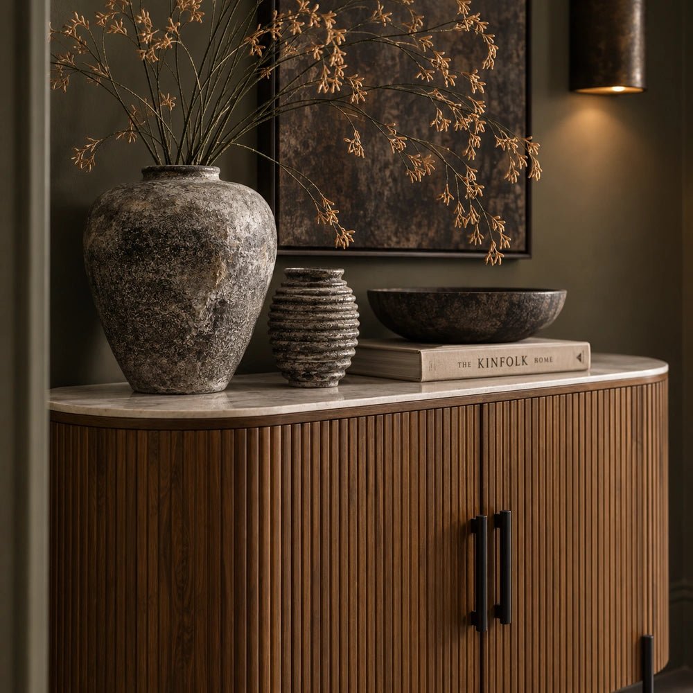 Warm wooden Hempton cabinet with vertical ribbed detailing and dark handles, styled with sculptural vases, books and decorative objects in a moody interior.
