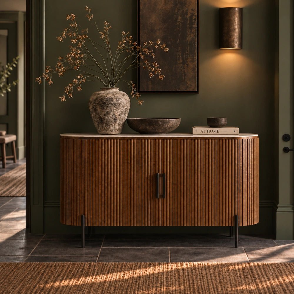 Warm wooden Hempton cabinet with vertical ribbed detailing and dark handles, shown close up with sculptural vases, books and decorative objects in a moody interior.