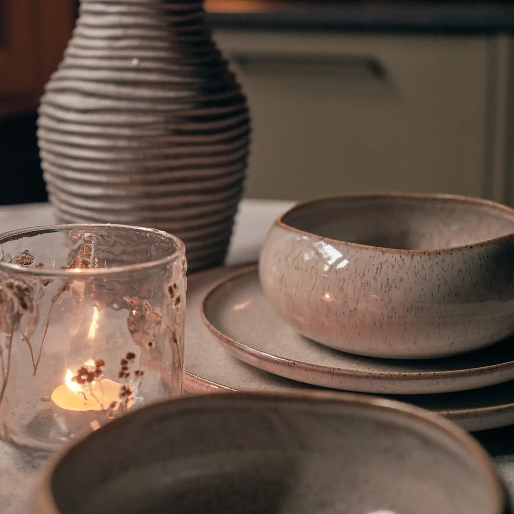 Hollister bowl displays its rounded shape and glossy speckled taupe glaze beside a candleholder, with a ribbed vase behind it on the dining table.