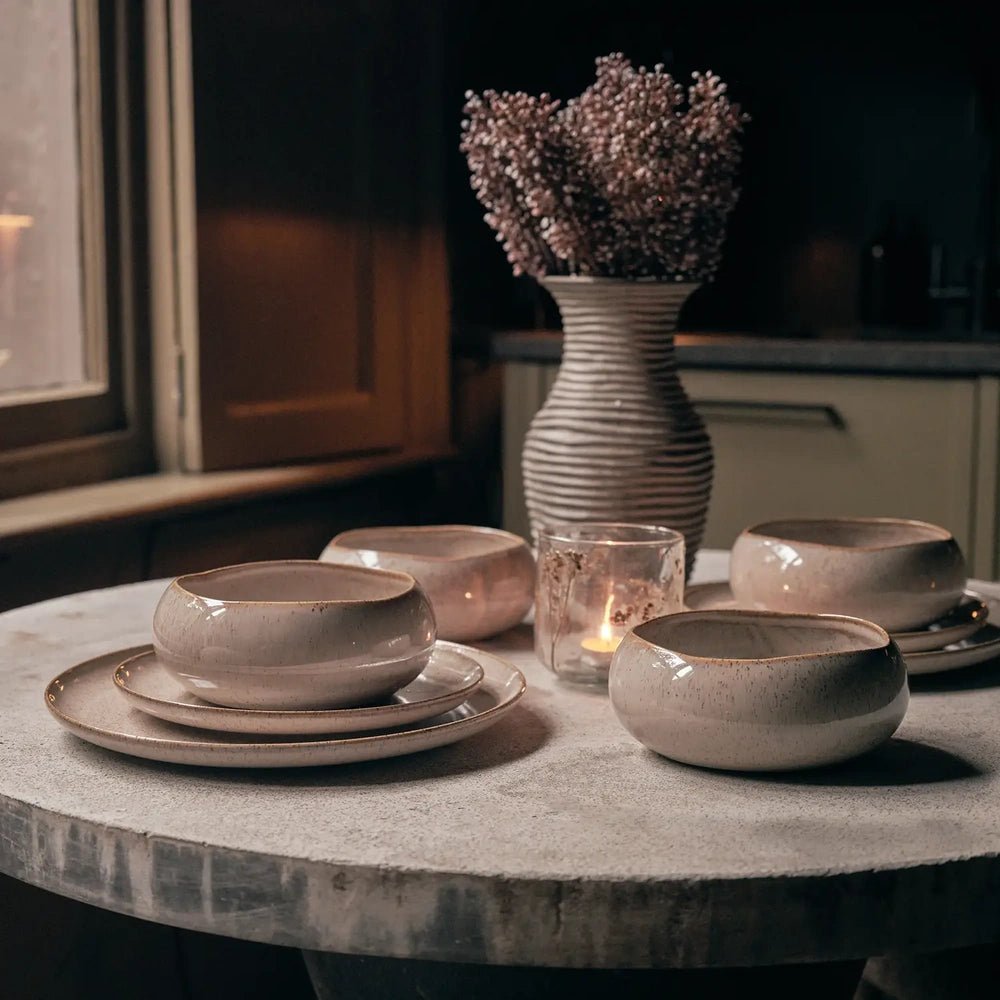 Hollister dinnerware in muted taupe is arranged on a round stone dining table with glowing candles, a ribbed vase and dried flowers beside a window.