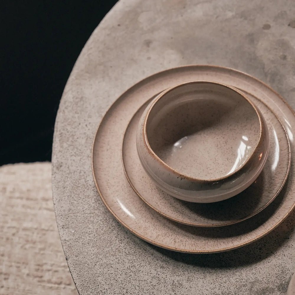 Hollister taupe plates and bowls are stacked on a pale stone tabletop, showing their rounded shapes, speckled glaze and softly irregular edges from above.