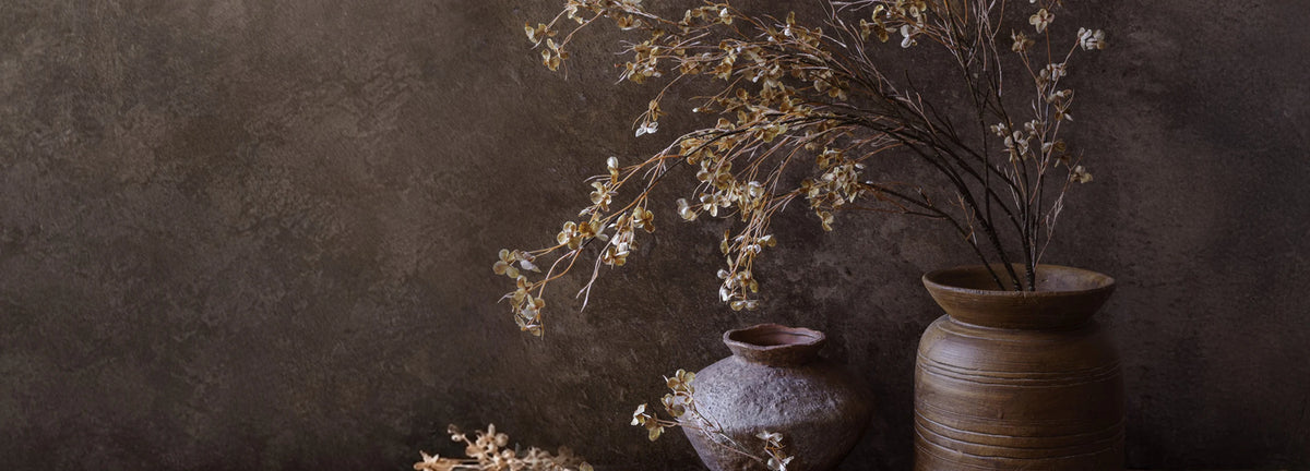 Cream and beige botanical branches arranged in two textured brown vases, with loose stems across a rustic wooden table against a dark textured wall.