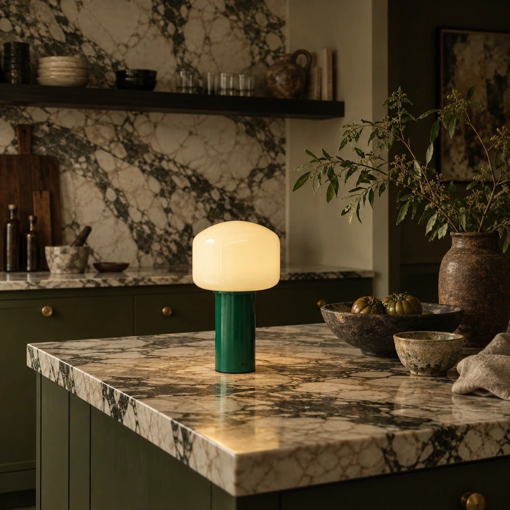 Green Hudson portable table lamp with softly glowing rounded shade, positioned on a dramatic marble kitchen island among rustic ceramics and leafy branches.