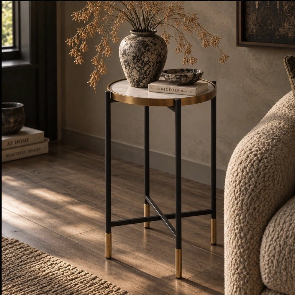 Huxton Side Table with slim black frame and warm metallic top, styled beside a textured armchair with books, a mottled vase and dried stems.