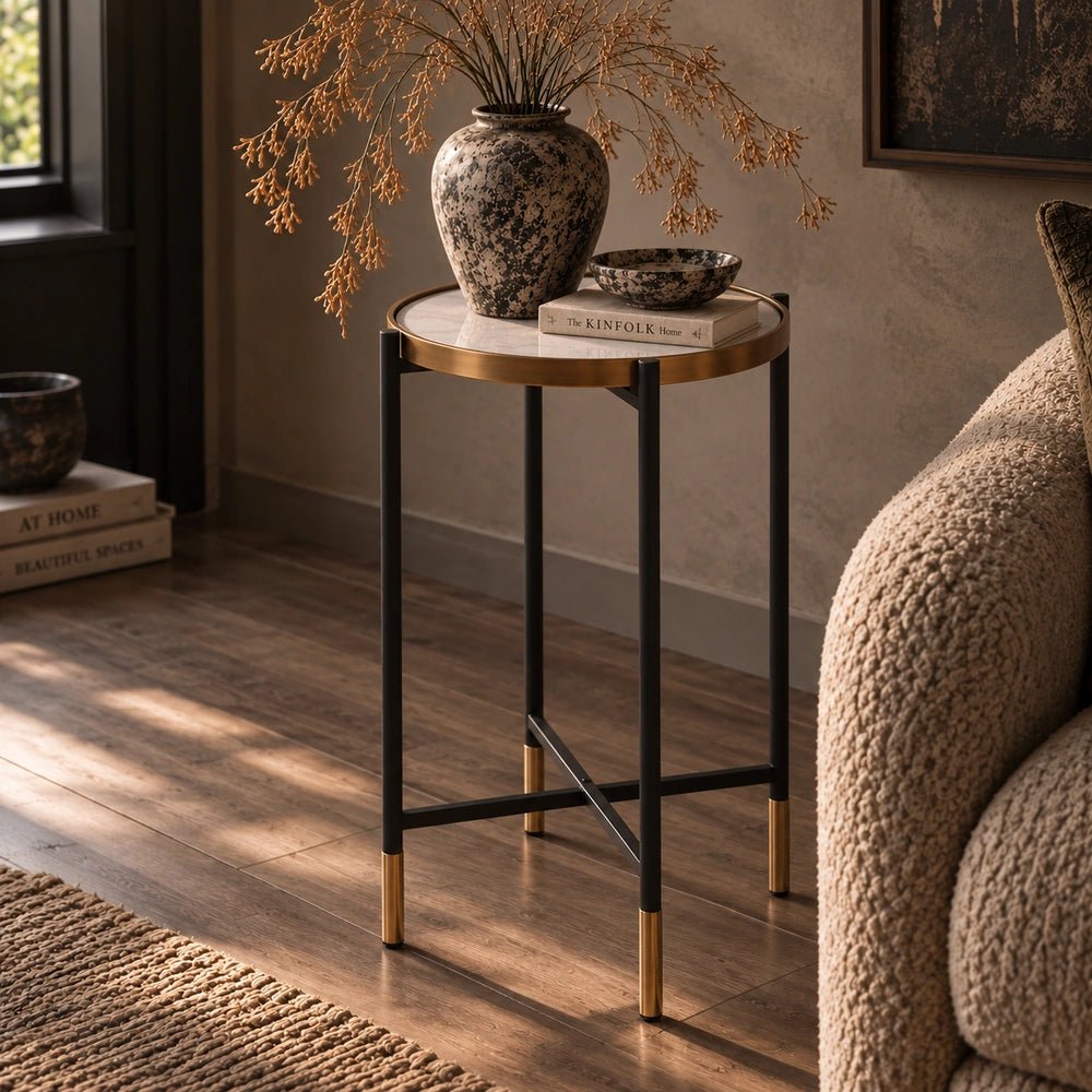 Huxton Side Table with round pale top and dark slim frame, styled beside a textured armchair with a large vase, bowl and stacked books.