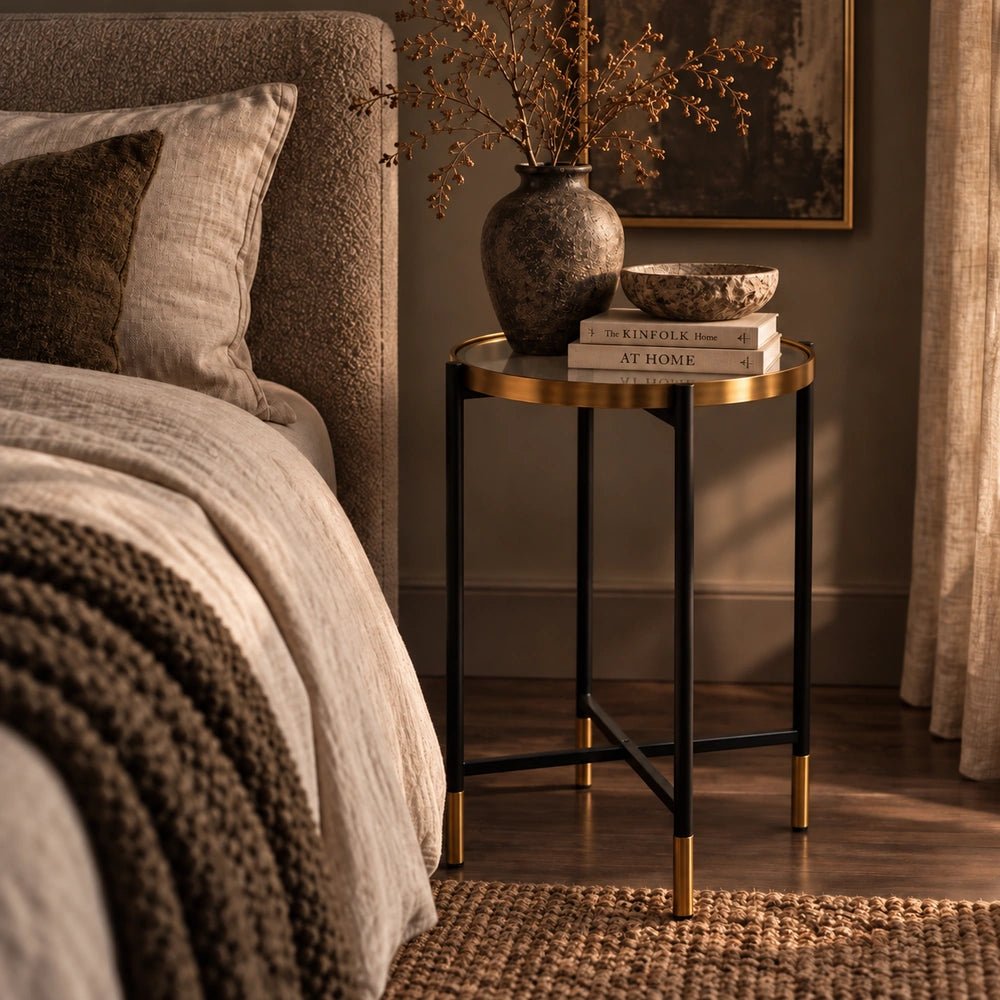 Huxton Side Table with slim dark frame and warm metallic top, styled beside a bed with books, a dark vase and soft neutral bedding.
