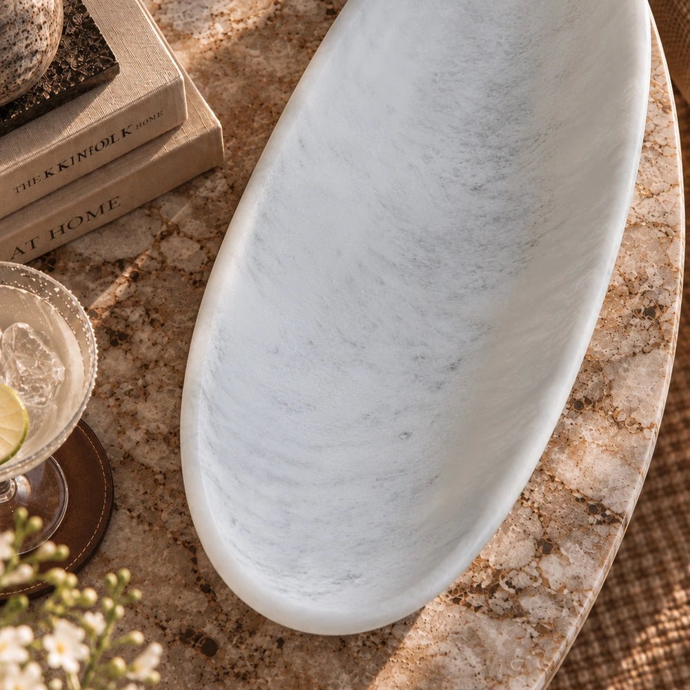 Irwin Resin Dish in soft white with an elongated oval shape and subtle marbled texture, shown from above on a stone table beside books and glassware.