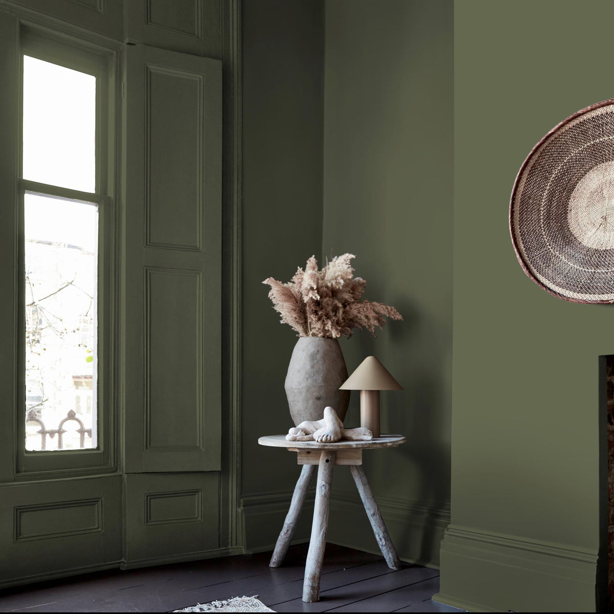 A wooden side table with a vase and table lamp in the corner of a room next to a window, with the walls painted in a dark green paint colour.