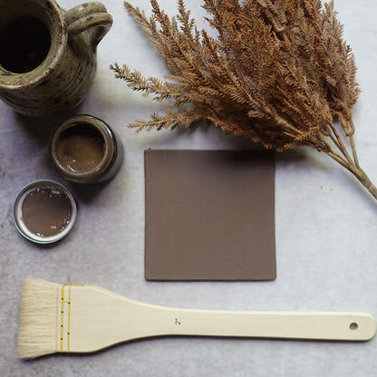 A warm brown painted tile next to a brush and an open sample pot