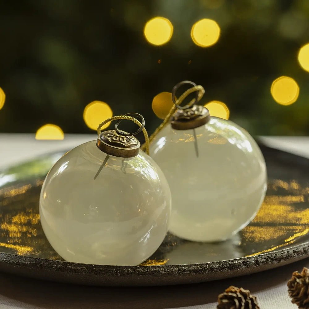 Kendal medium opal bauble with a rounded pearl white finish and hanging loop rests on a decorative plate beside another ornament against warm Christmas lights.