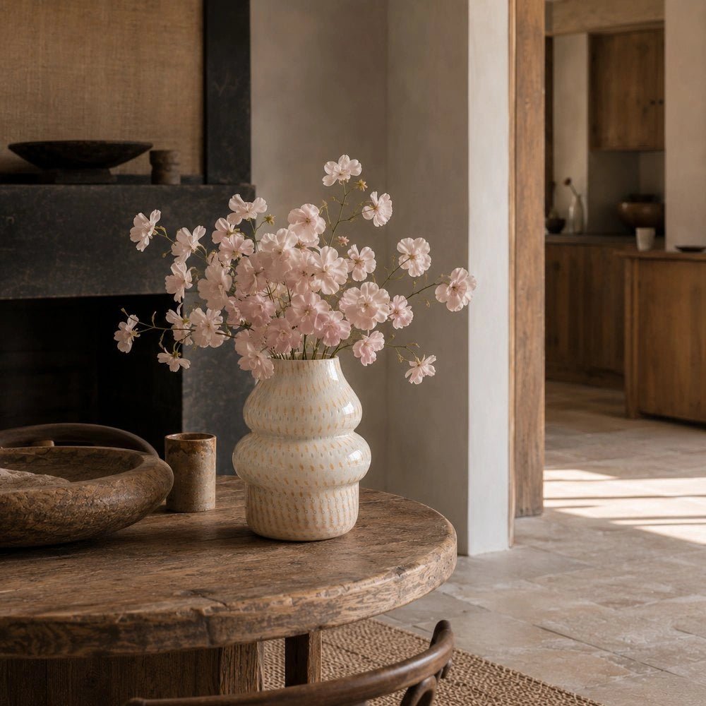 Lagor Glass Vase with rounded textured cream silhouette, filled with pale pink blossom stems and styled on a rustic round table in a warm neutral interior.