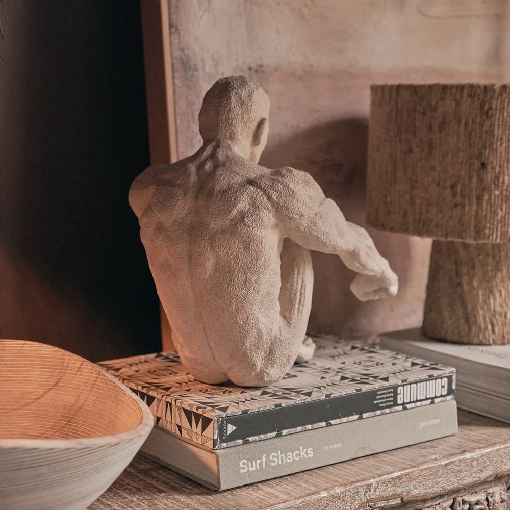 Lawrence Figurative Sculpture depicts a pale seated figure with a textured surface, displayed on stacked books beside sculptural accessories on a wooden console.