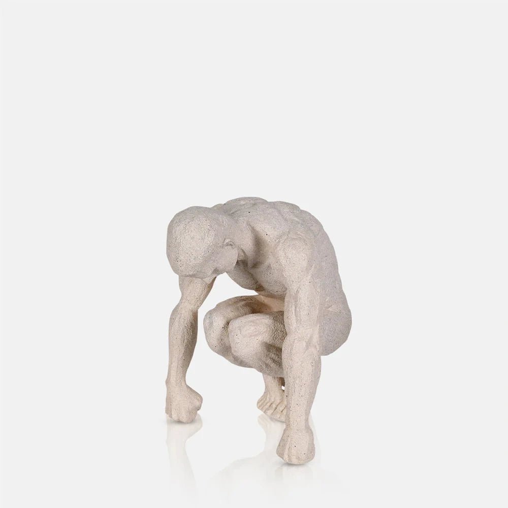 Lawrence Figurative Sculpture depicts a pale seated figure with a bowed head, bent knees and elongated arms, finished with a softly textured sculptural surface.