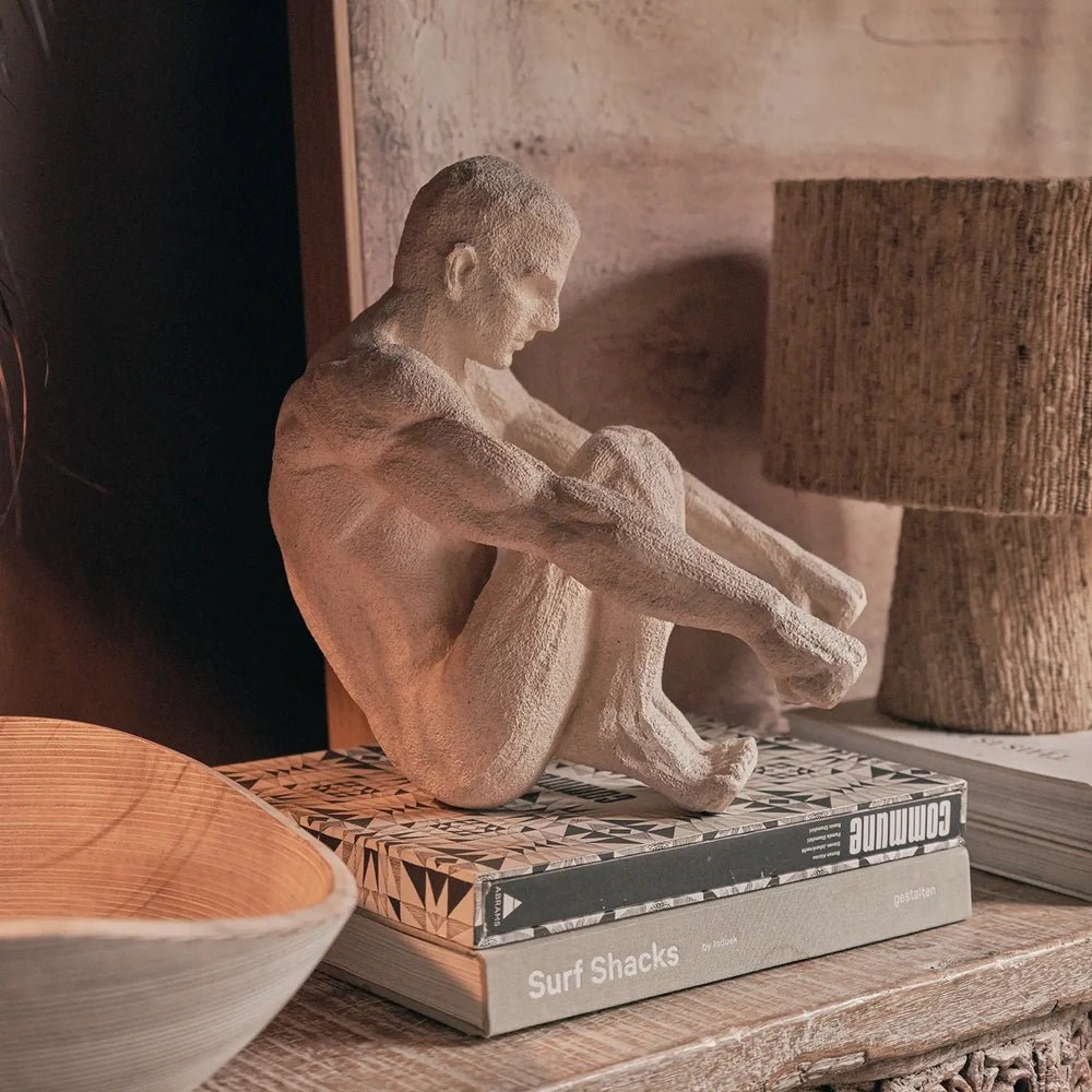 Lawrence Figurative Sculpture presents a pale seated figure in side view, with bent knees and textured contours, displayed on books beside a cylindrical lamp.