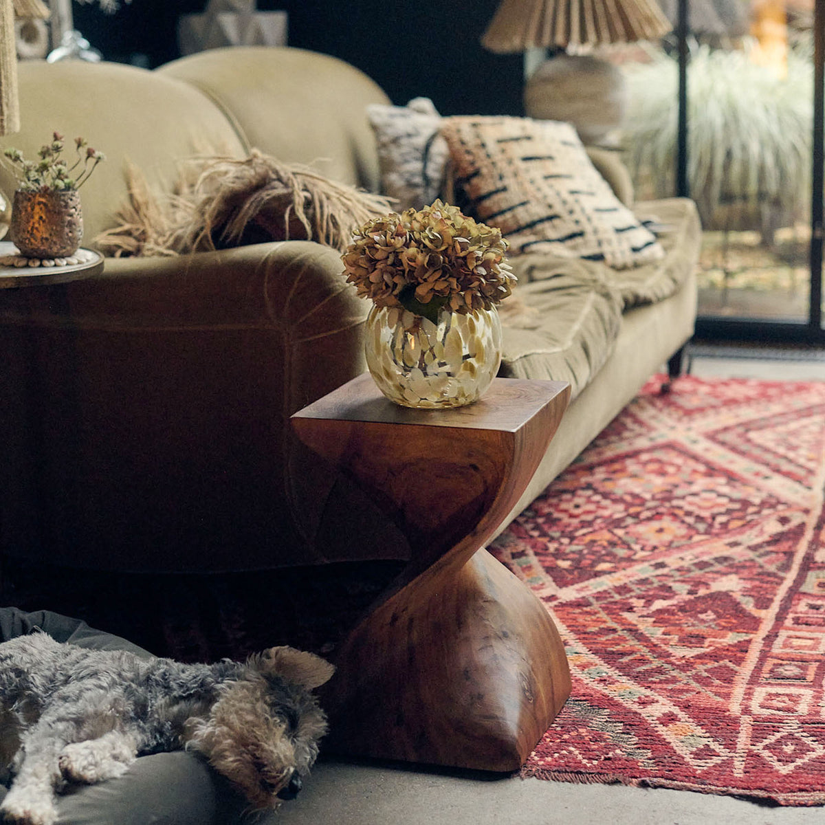 A modern wooden side table with a vase of faux flowers in front of a green sofa.