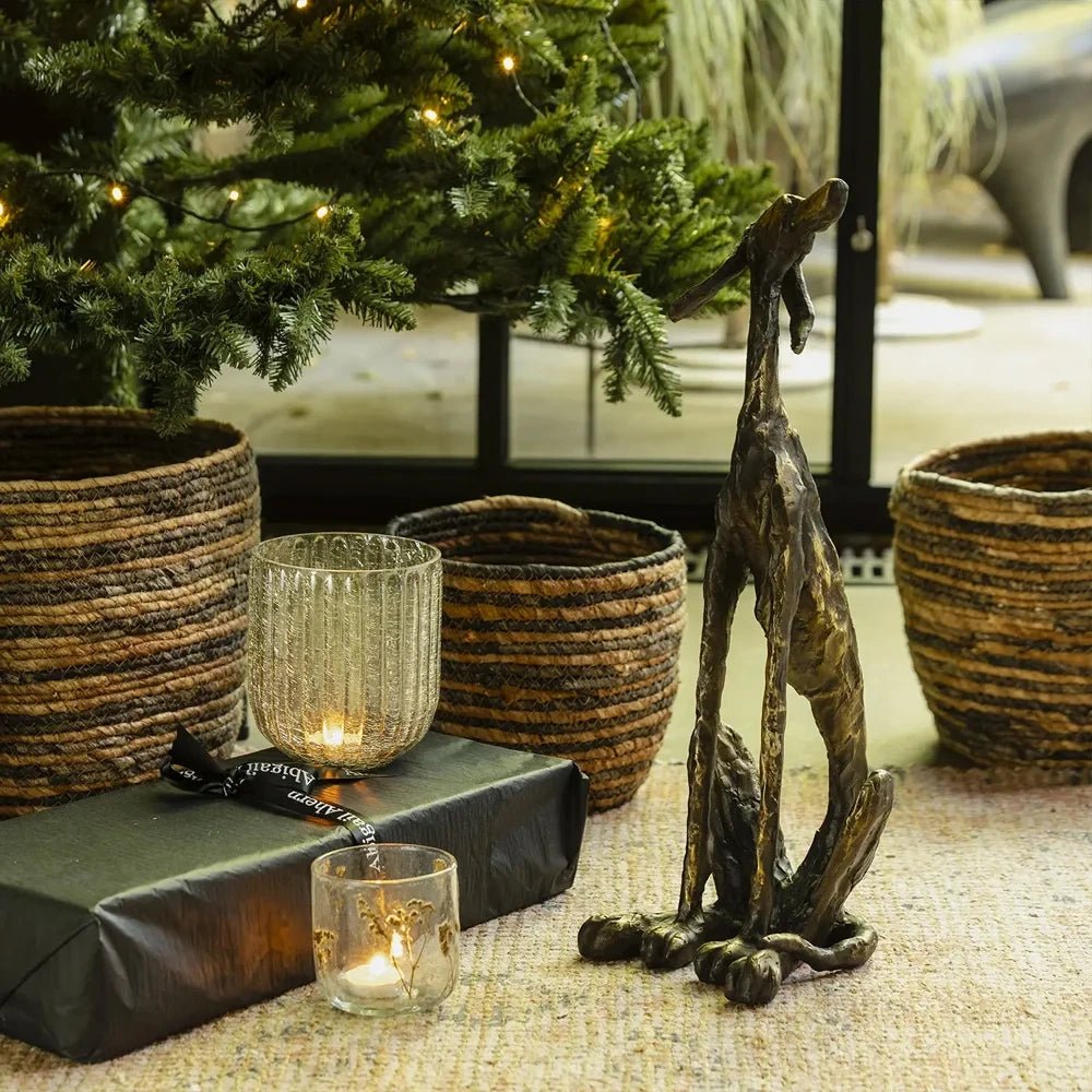 The Linley glass candle holder joins glowing votives beside black wrapped gifts, striped woven baskets and a tall hare sculpture under illuminated Christmas branches.
