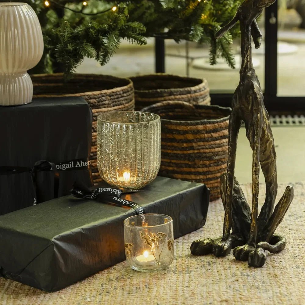 The Linley glass candle holder sits among glowing glass votives, black wrapped gifts and woven baskets beside a tall hare sculpture beneath a Christmas tree.
