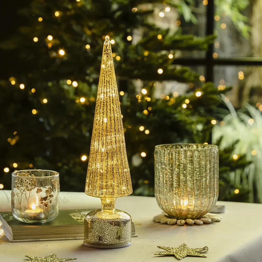 The Linley glass candle holder appears in a gold-toned festive display with glowing votives, an illuminated cone-shaped tree, scattered stars and Christmas lights behind.