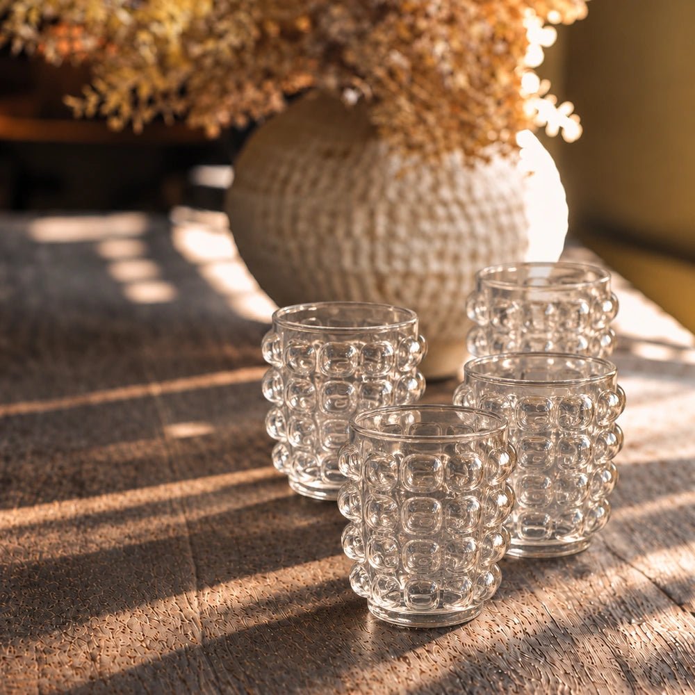 Four Lorien Bobble Glass tumblers with clear raised bobble texture, grouped on a sunlit wooden table beside a textured vase and warm neutral decorative styling.