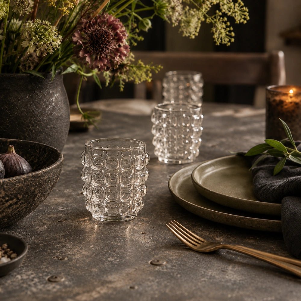 Two Lorien Bobble Glass tumblers with clear raised bobble texture, styled on a dining table among dark plates, foliage and softly layered neutral tableware.