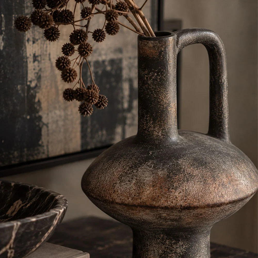 Lovina vase with a dark mottled finish, rounded body, narrow neck and tall loop handle holds dried branches beside a decorative bowl on a console.