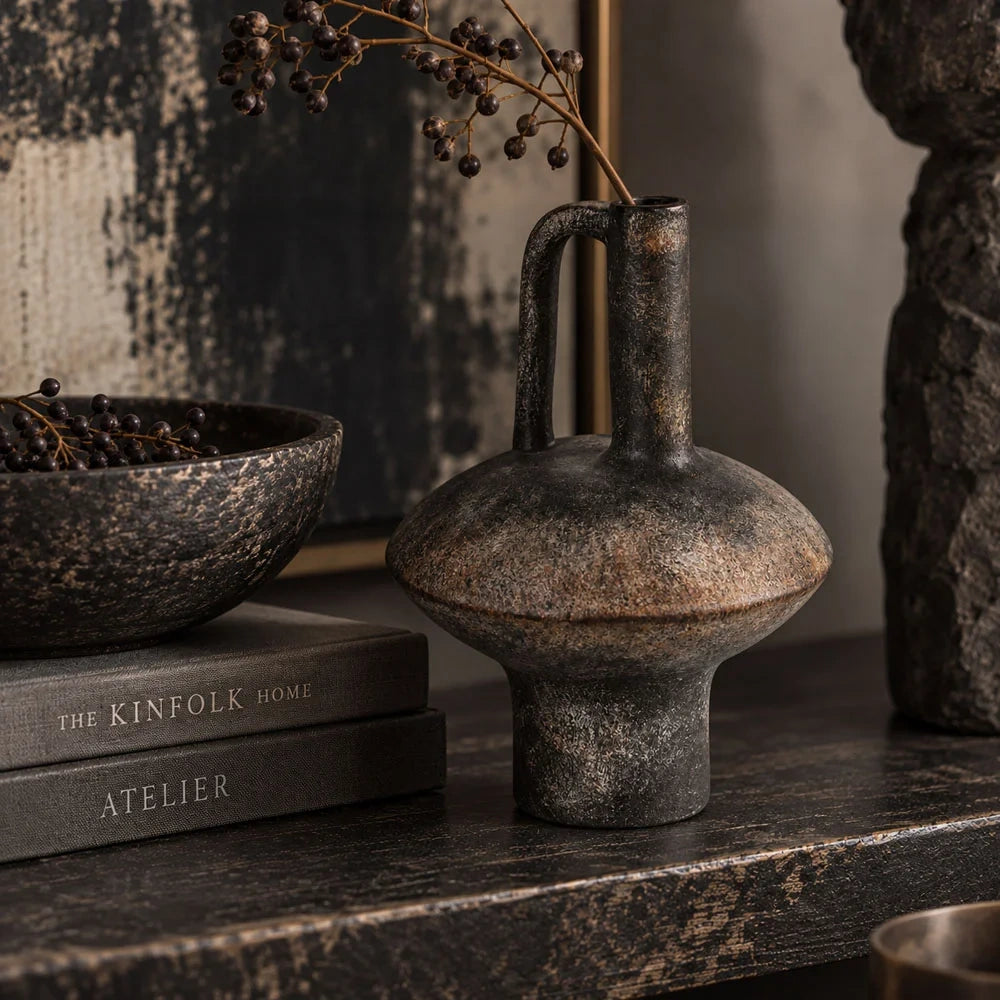 Lovina vase with a dark mottled finish, rounded body and tall loop handle stands beside stacked books and a decorative bowl on a dark console.