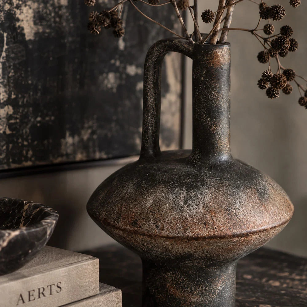 Lovina vase shown close up, highlighting its tall loop handle, narrow neck and rounded dark body with mottled bronze brown tones beneath dried branches.