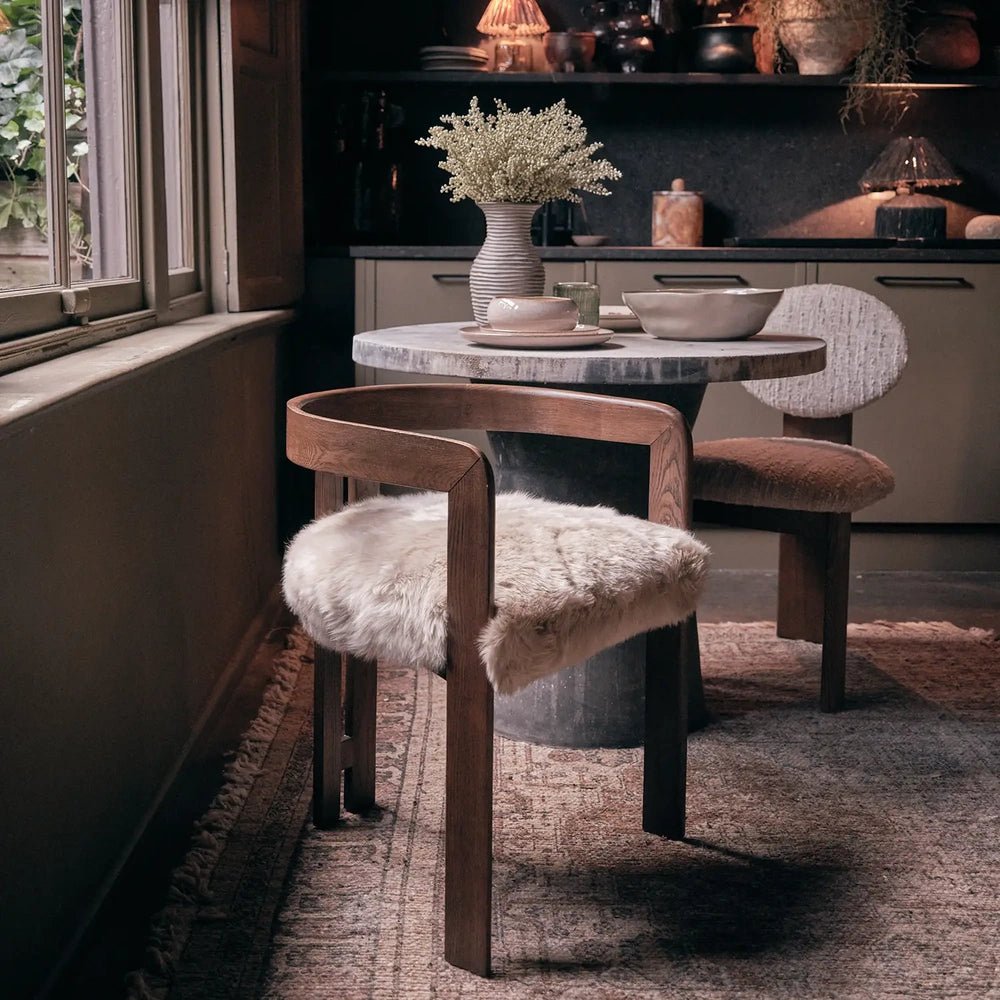 Loxford Sheepskin Dining Chair features a pale shaggy seat within a warm wooden frame, positioned beside a dark round table in a styled dining room.