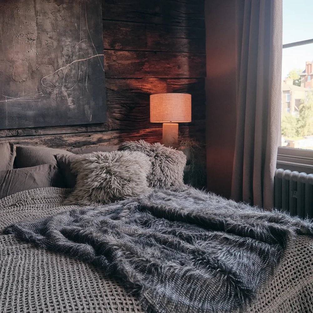 Malton Throw spreads across the foot of a bed, its long grey fibres contrasting with smooth bedding beneath a warm lamp and tall bedroom window.