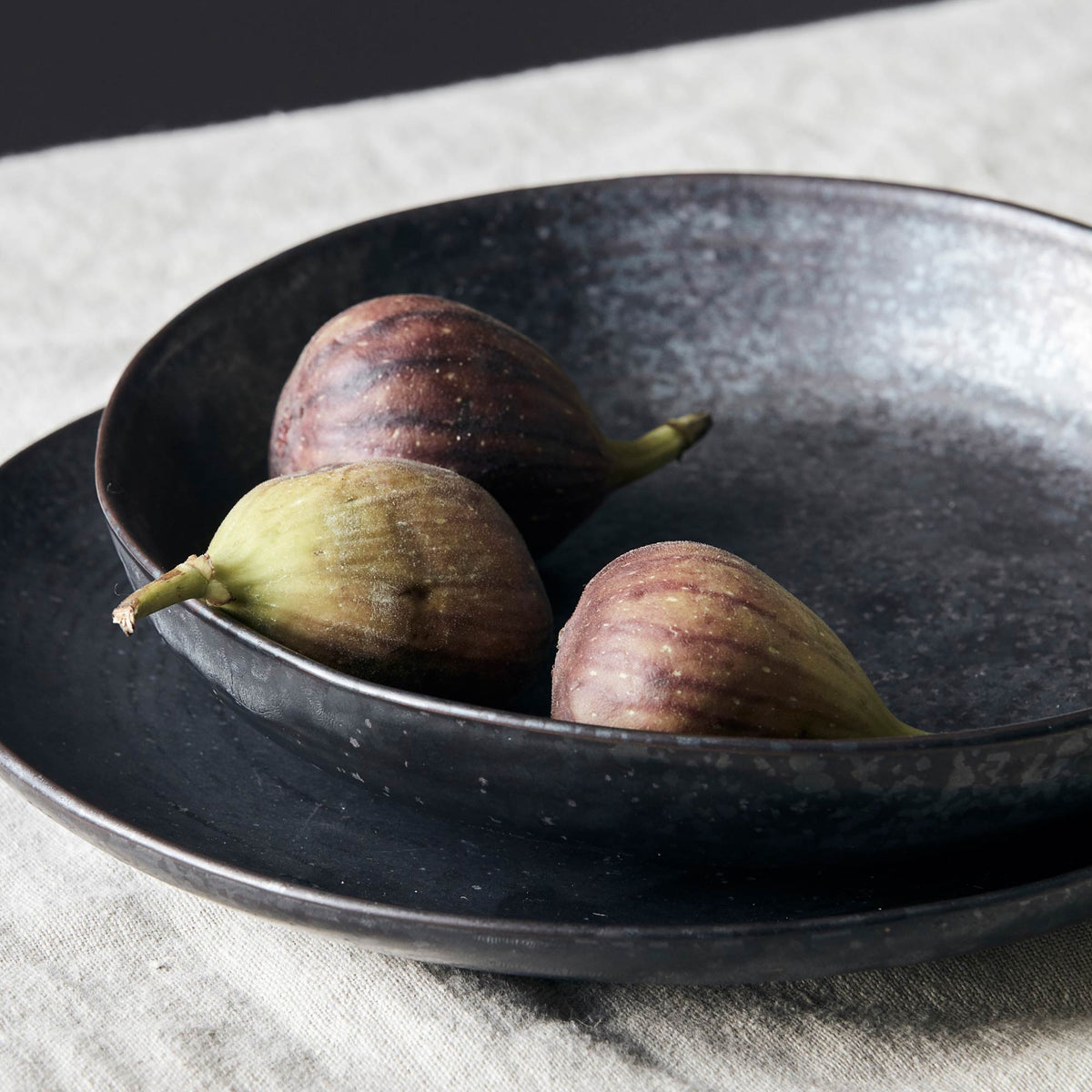 The Marple Stoneware Bowl Charcoal, a wide shallow stoneware bowl in an inky black colour, containing three figs.