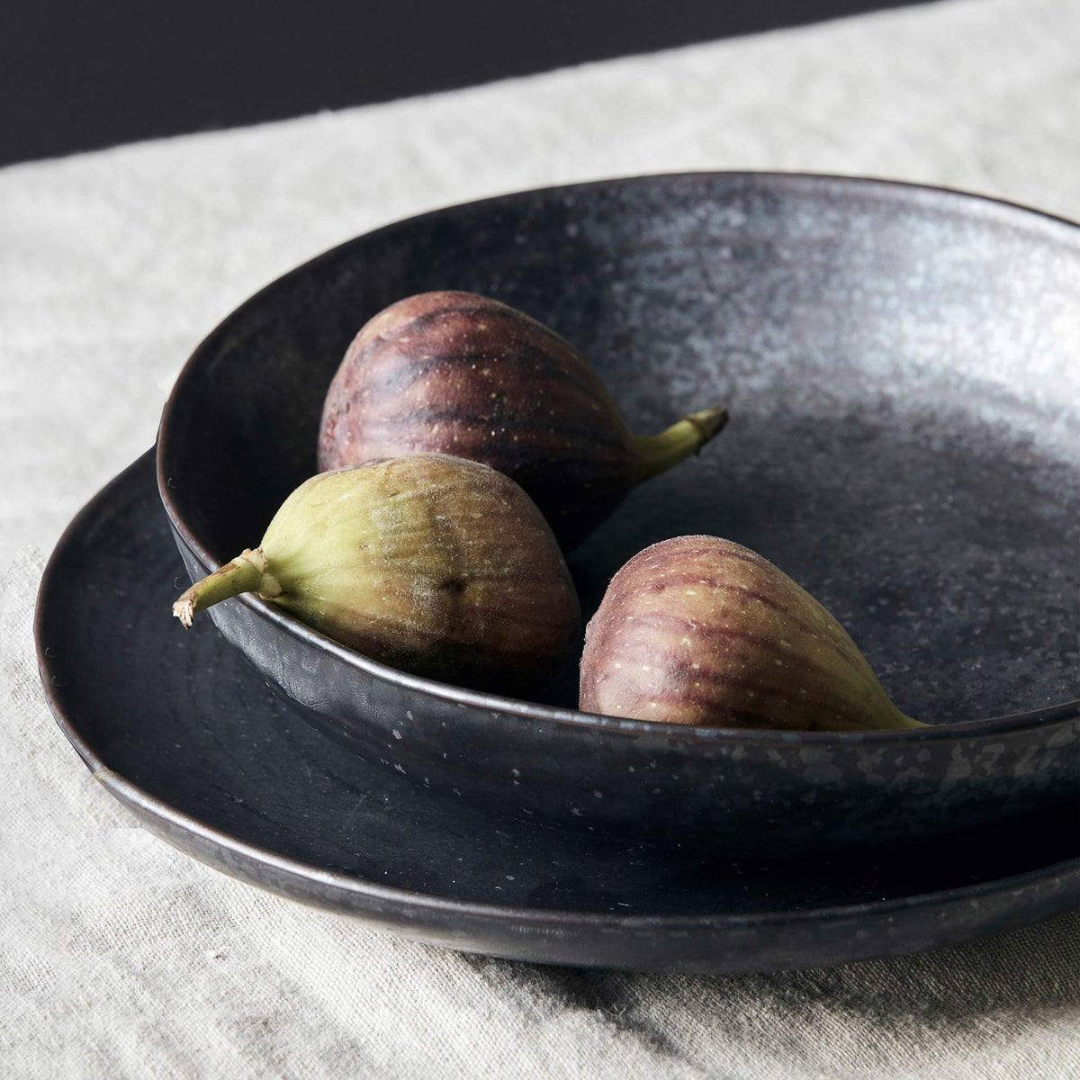The Marple Stoneware Bowl Charcoal, a wide shallow stoneware bowl in an inky black colour, containing three figs.