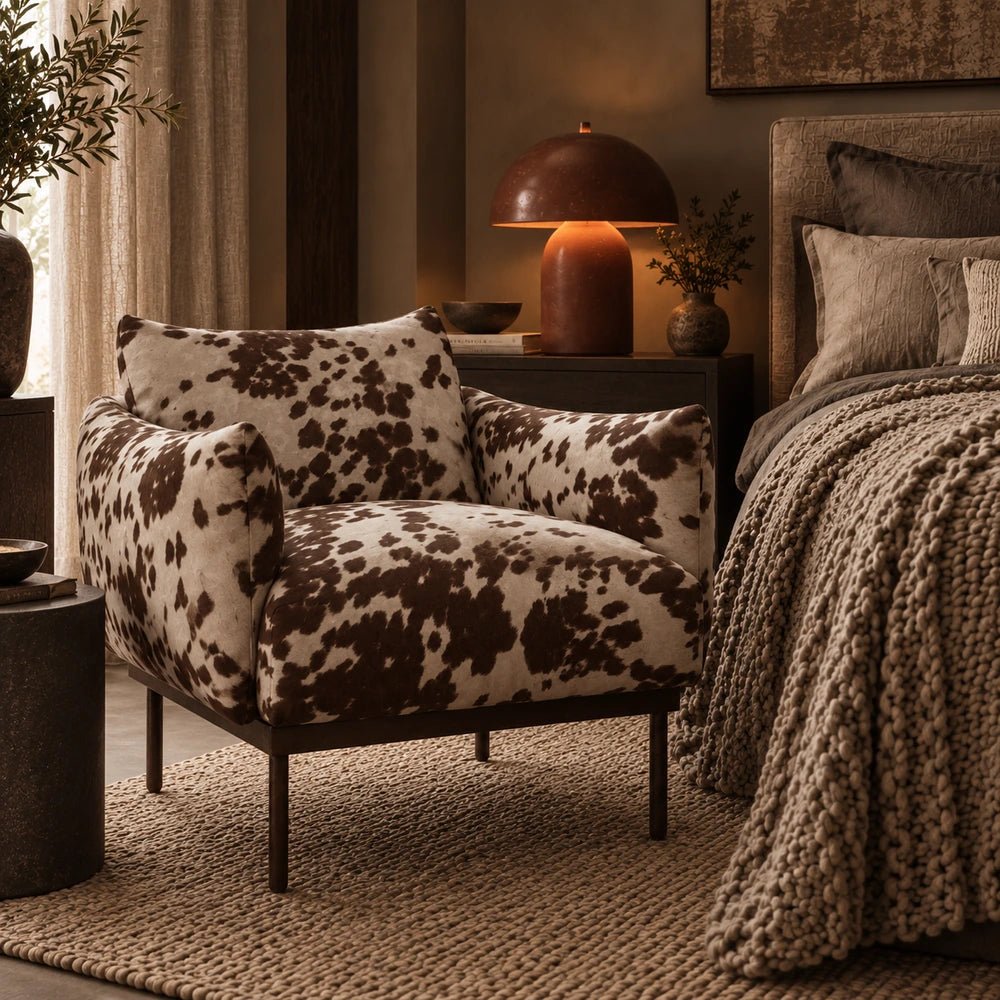 Melvin Armchair in a warm bedroom, showing its brown and cream cowhide-style pattern, cushioned arms and seat beside a textured bed and table lamp.