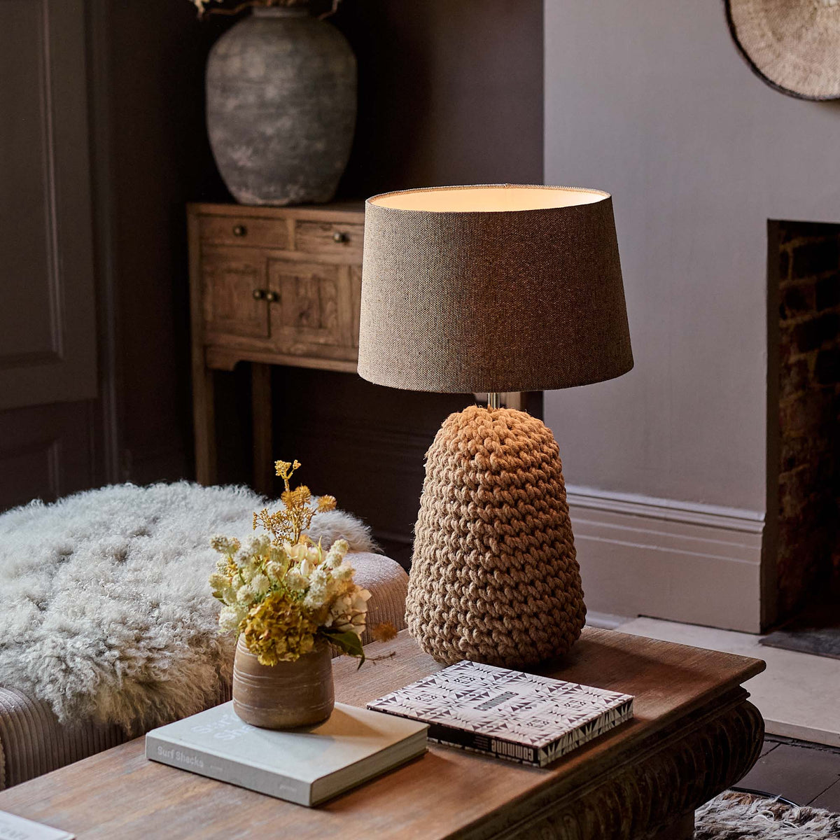The Milton Table Lamp, a table lamp with a chunky knotted base in warm brown and a fabric drum shade, on a coffee table.