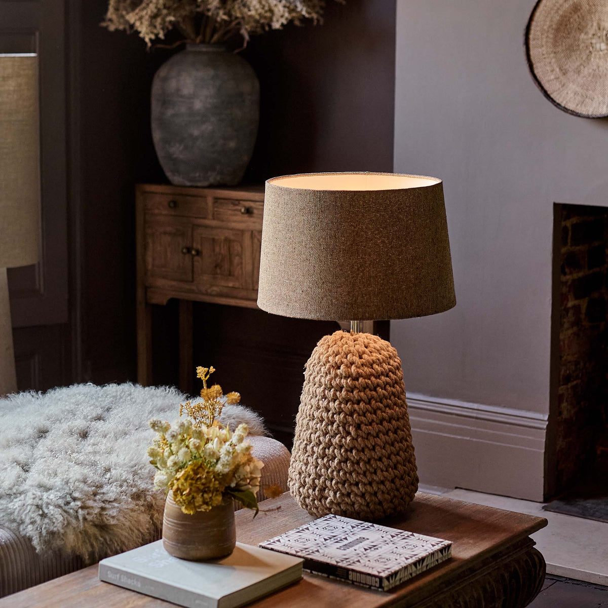 The Milton Table Lamp, a table lamp with a chunky knotted base in warm brown and a fabric drum shade, on a coffee table.