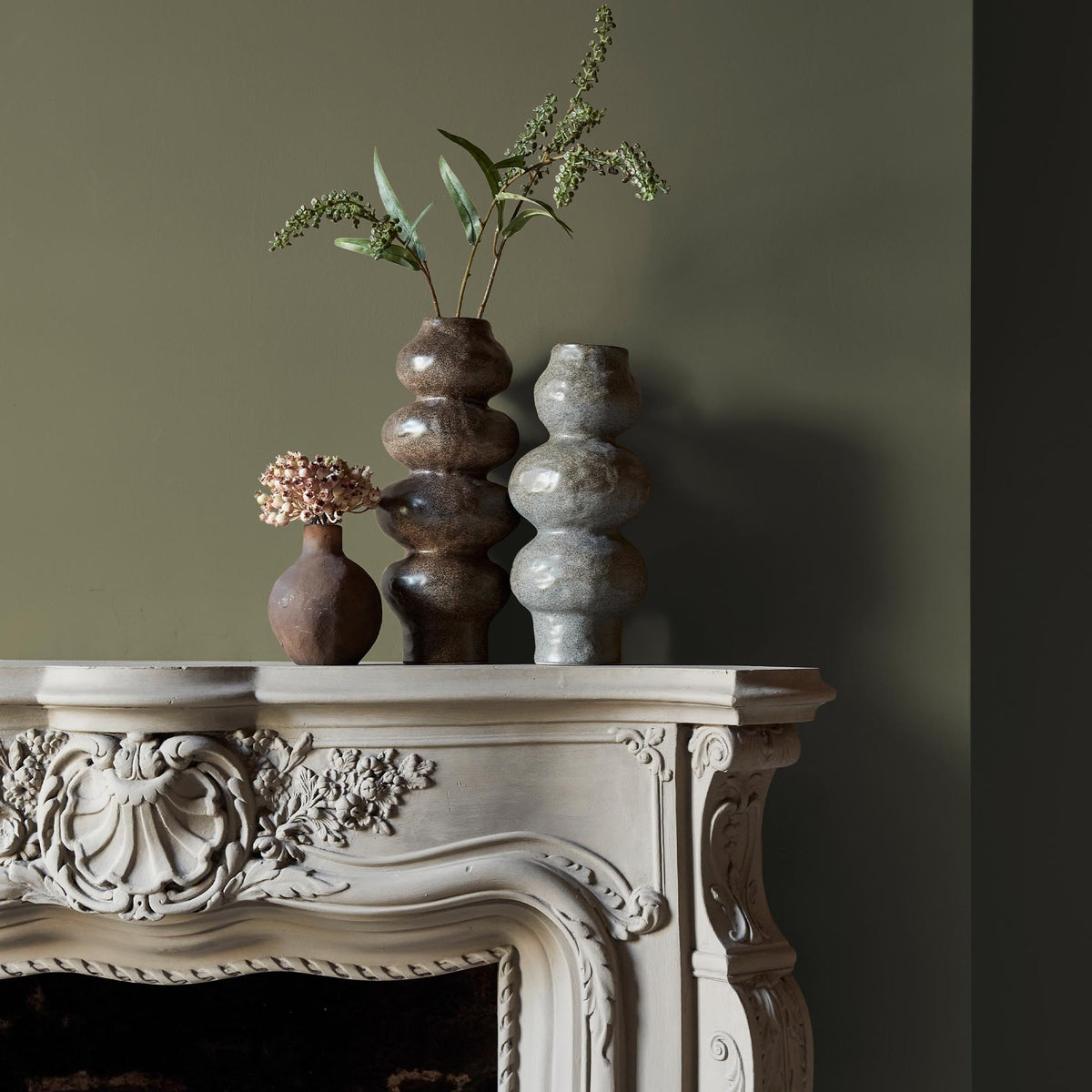 Three ceramic vases sitting on an intricate fireplace mantle in front of a wall in a dark, sage green paint colour.