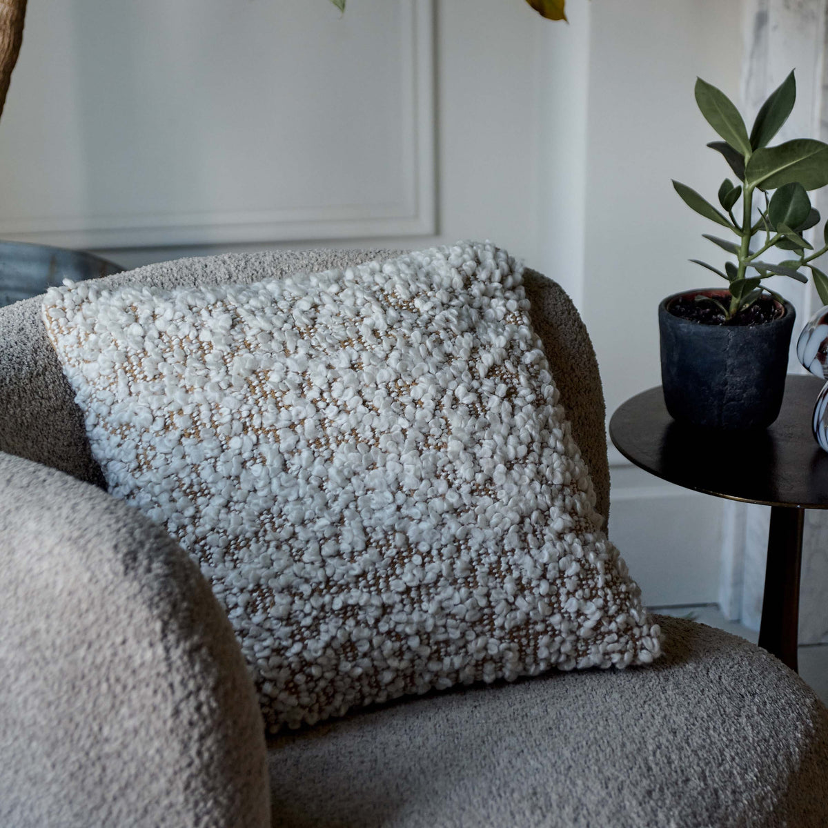 The Hallet Boucle Cushion, a square cushion in warm brown with cream boucle accents placed on a curved cream boucle armchair.