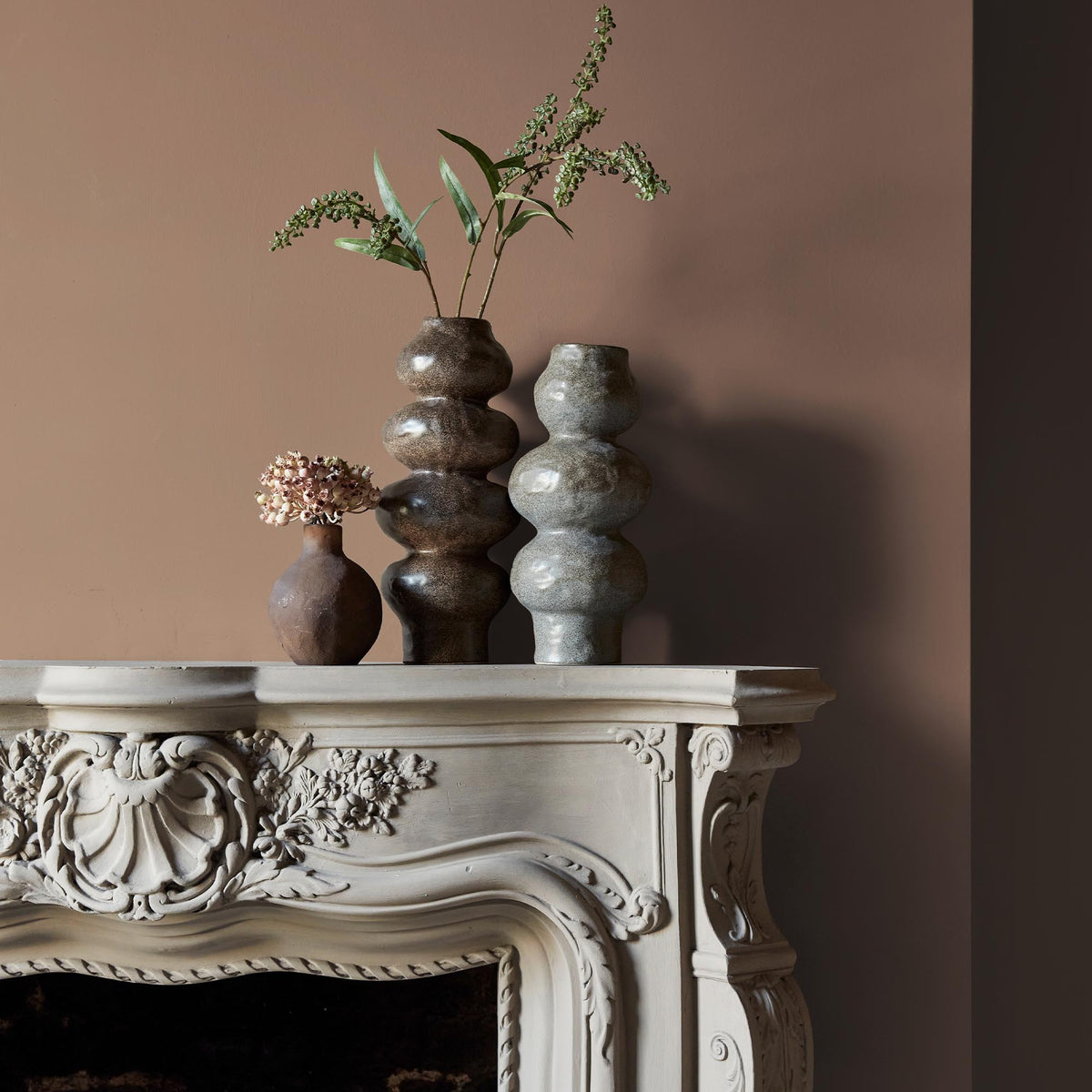 Three ceramic vases sitting on an intricate fireplace mantle in front of a wall in a dusky pink paint colour with a warm orange undertone.