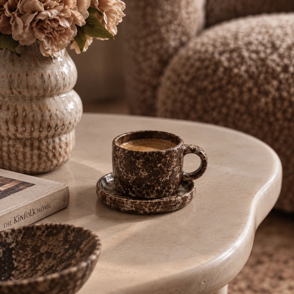 Norman Cup and Saucer, Mocha filled with coffee on a pale rounded table, showing speckled brown ceramic beside flowers, a book and bowl.