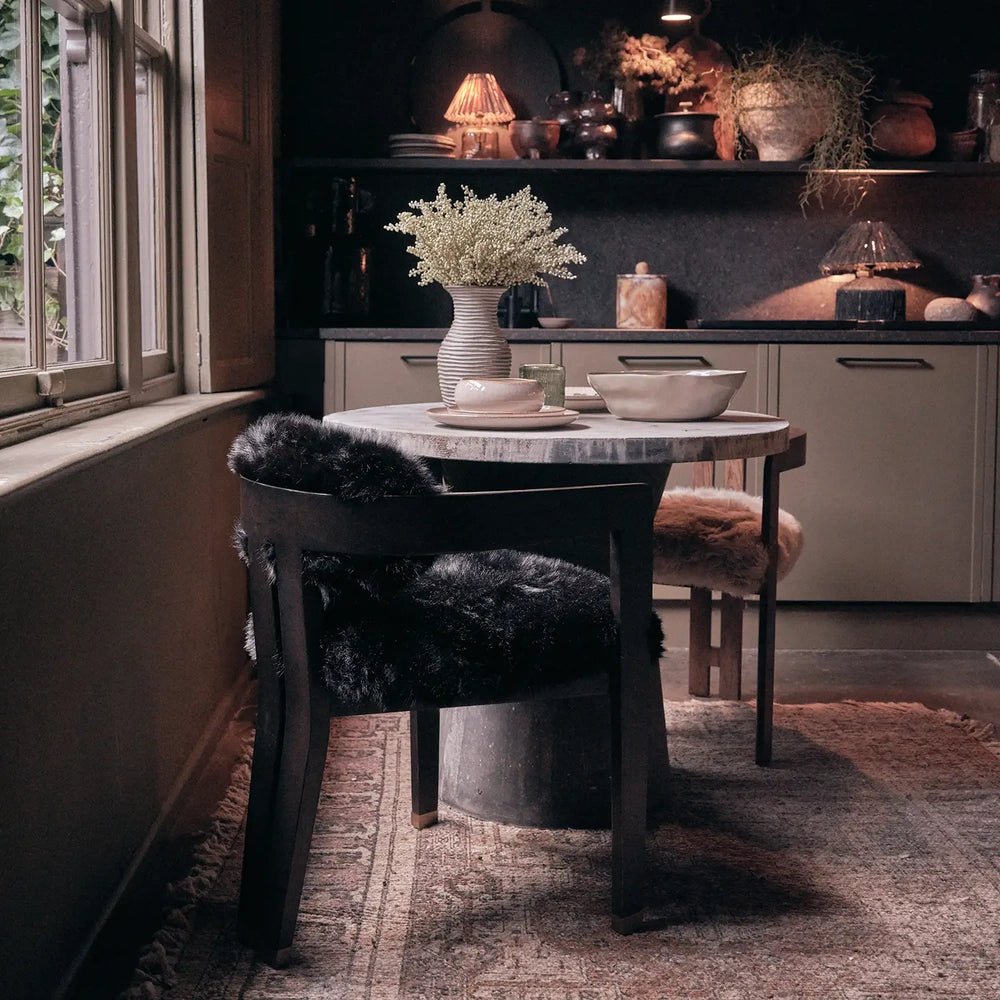 Norwyn Faux Fur Dining Chair has a dark wooden frame and black textured upholstery, shown beside a round dining table near a window and fireplace.