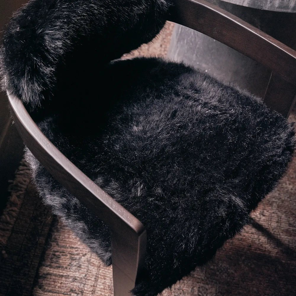 Norwyn Faux Fur Dining Chair shows its shaggy black upholstery and dark wooden armrest in close-up, highlighting the deep seat and contrasting smooth frame.